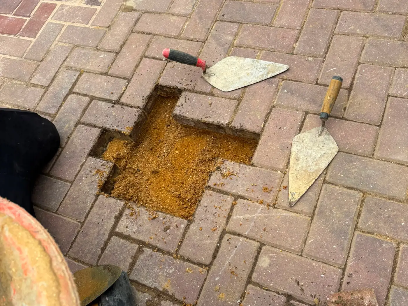 Close up of block paving being laid, with a trowel, lump hammer and one block lifted to show the sand bed beneath