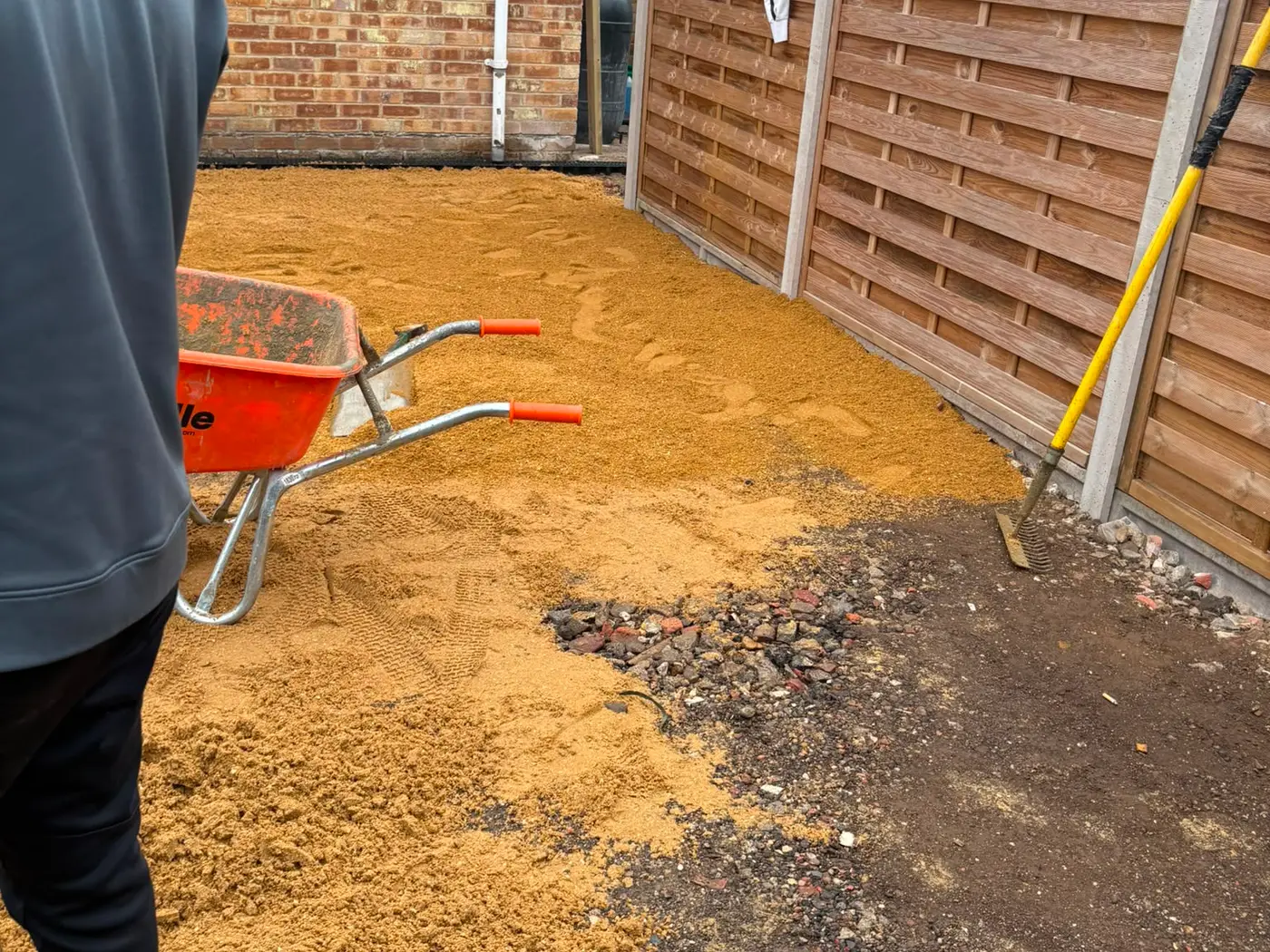 One of the team barrowing sand across the prepared base of a driveway beside a house